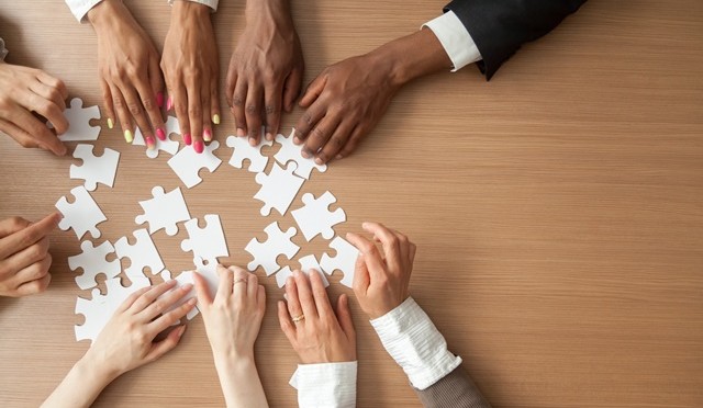 Hands of multi-ethnic team assembling jigsaw puzzle, multiracial group of black and white people joining pieces at desk, successful teamwork concept, help and support in business, close up top view